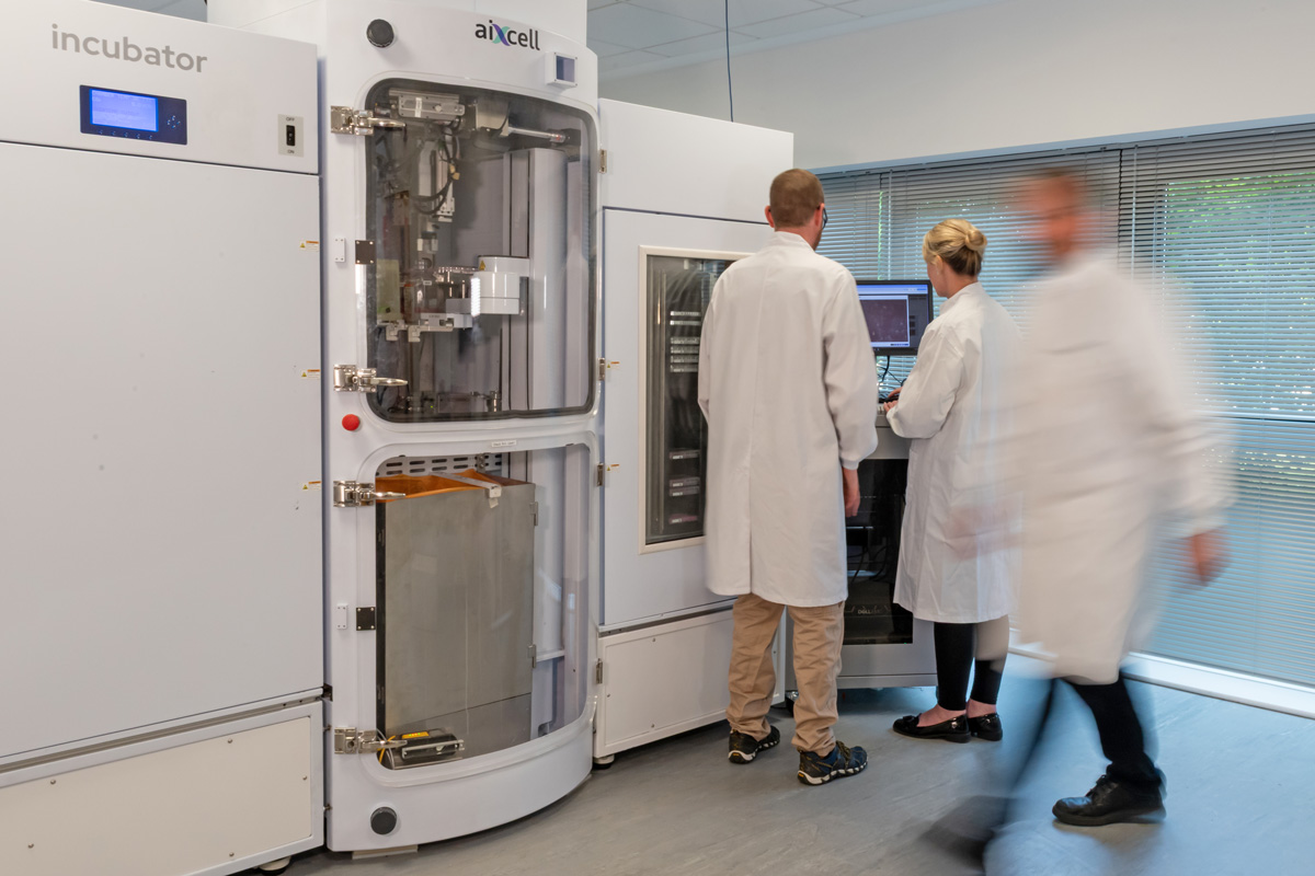 Three researchers discussing autonomous cell culture system UI; one blurred, walking past.